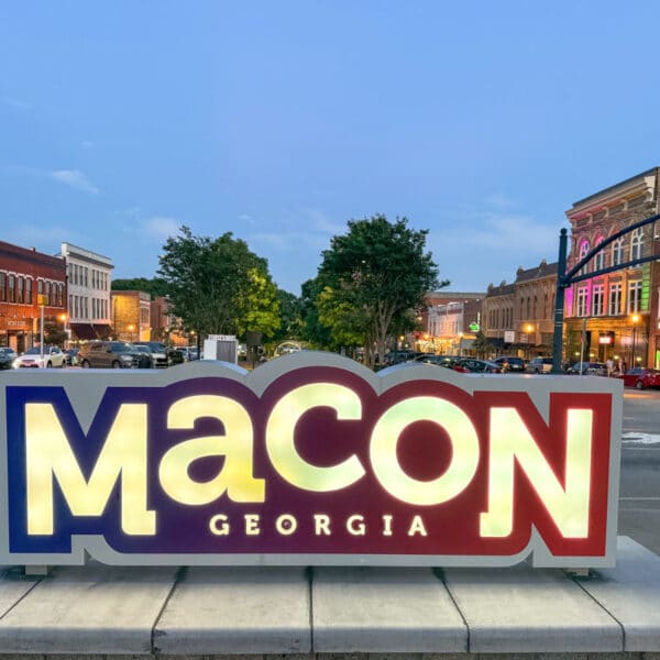 Illuminated sign reading “Macon Georgia” stands on a street corner, with historic downtown buildings and streetlights visible in the background at dusk.