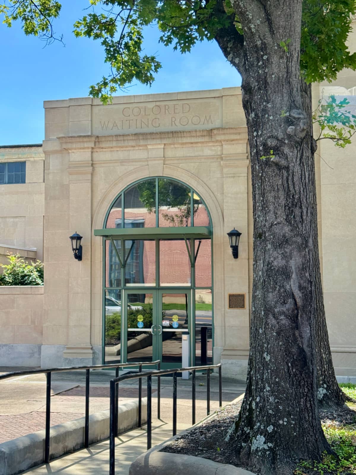 Stone building entrance labeled &ldquo;Colored Waiting Room&rdquo; above a large window and door, with a tree and ramp in the foreground.