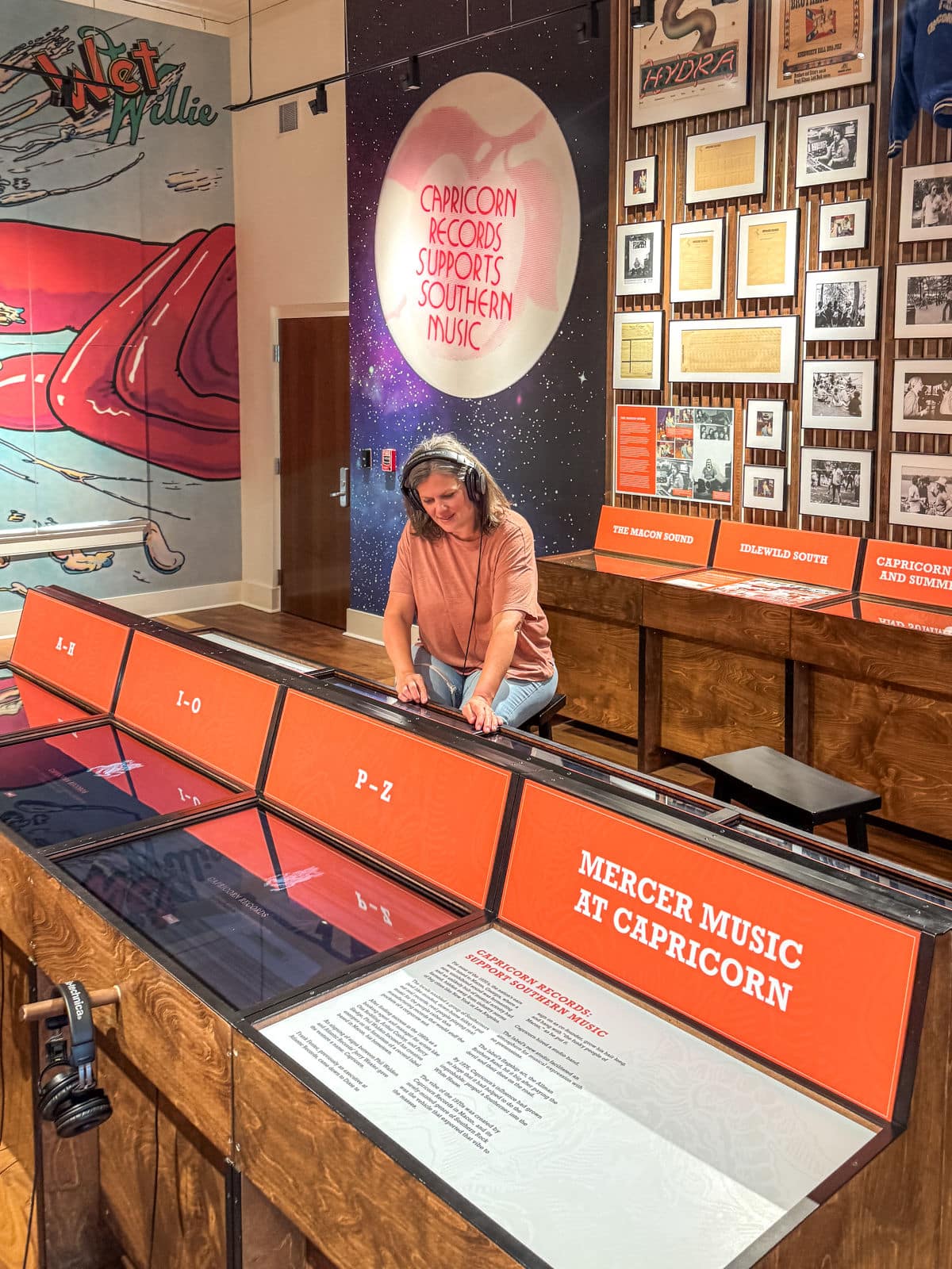A woman wearing headphones stands at an interactive music exhibit at Mercer Music at Capricorn, with displays and music-related wall art visible in the background.