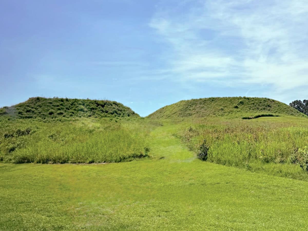 Two grassy, flat-topped earthen mounds stand under a blue sky, surrounded by green grass and sparse vegetation.