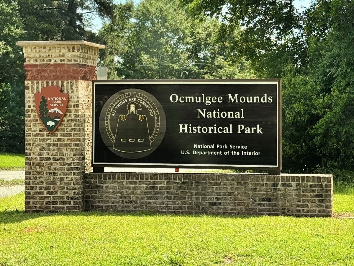 Brick pillar and sign at entrance to Ocmulgee Mounds National Historical Park, managed by the National Park Service, on a grassy area with trees in the background.