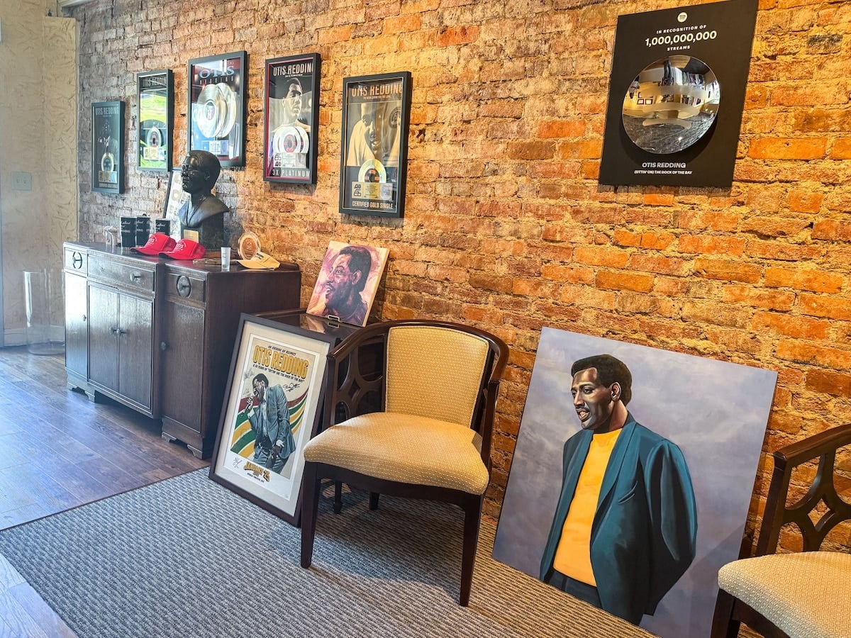 Room with exposed brick wall featuring framed posters, a bust, vinyl records, portraits, and two chairs on a carpeted floor.