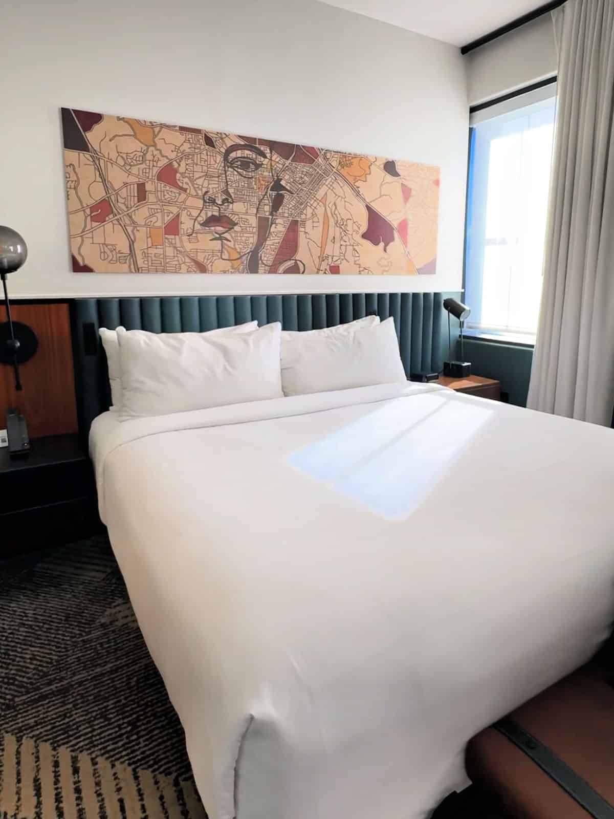 A neatly made bed with white linens, a map-themed artwork above the headboard, bedside tables, lamps, and a window with partially drawn curtains.