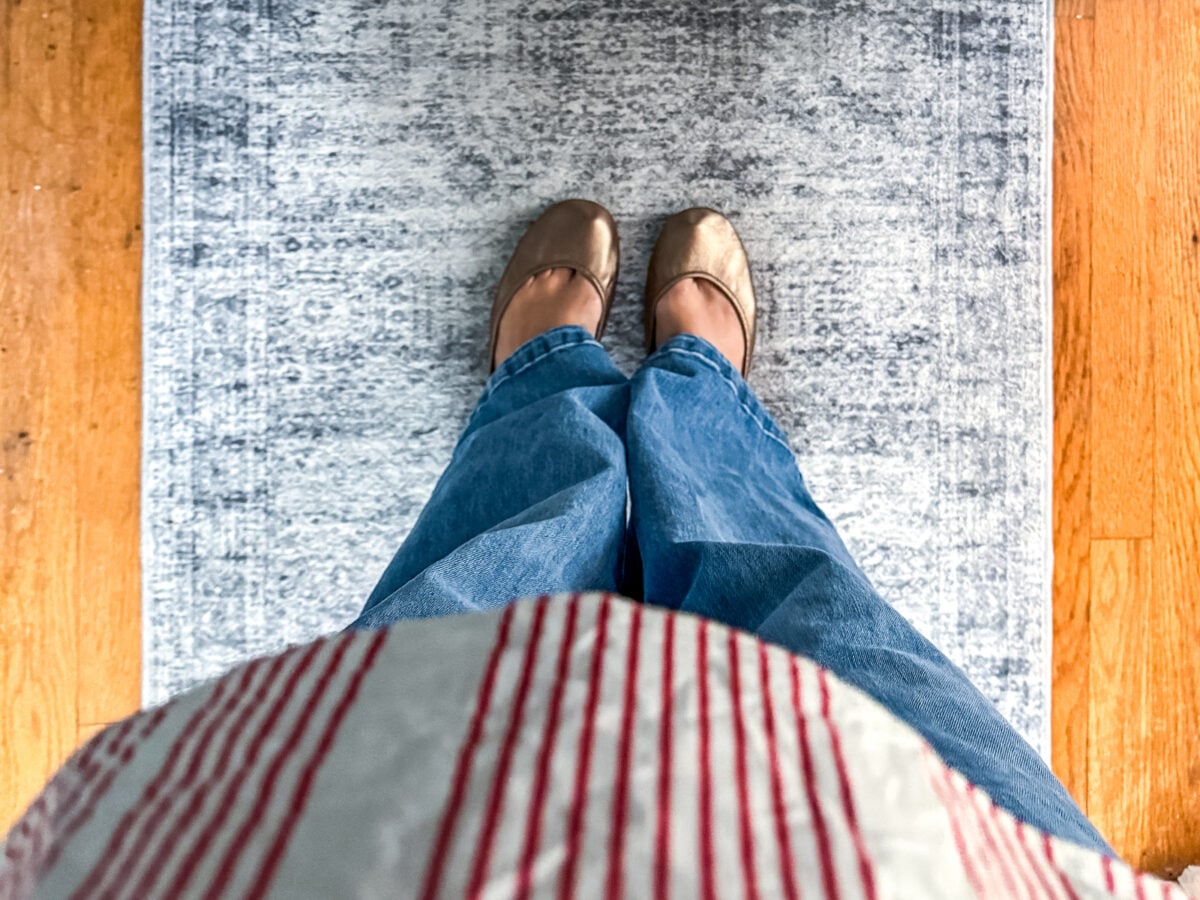 Person wearing blue jeans and gold flats stands on a light gray rug with a hardwood floor visible on both sides. Red and white striped shirt is partially visible at the bottom of the image.