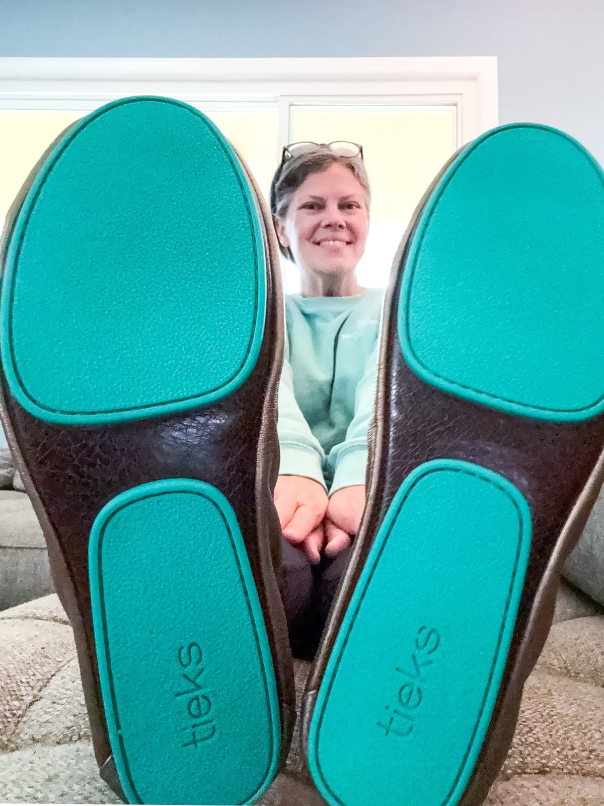 A woman sits on a couch, smiling, with her feet extended forward showing the turquoise soles of her brown Tieks flats.