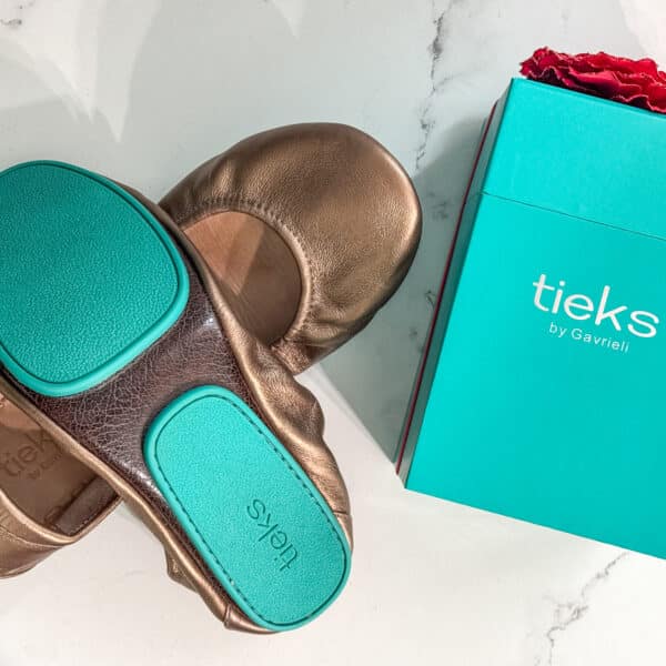 A pair of bronze Tieks ballet flats with teal soles next to a turquoise Tieks box with a red flower on top, on a white surface.