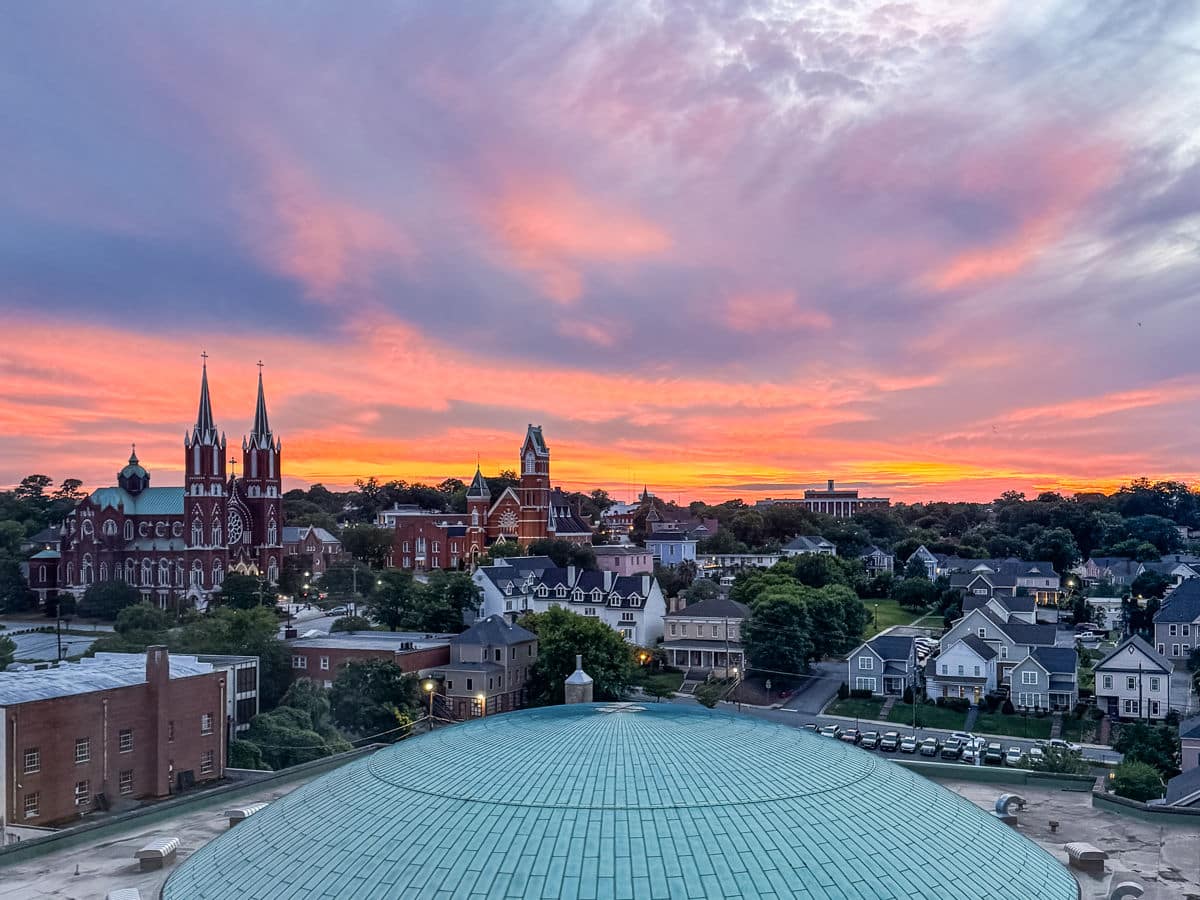 A cityscape featuring church spires, residential houses, and a green dome roof under a colorful sunset sky with orange, pink, and purple clouds.