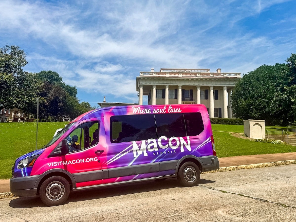 A colorful van labeled &ldquo;Macon Georgia&rdquo; is parked on a street in front of a large white mansion, with green lawns and trees under a blue sky.