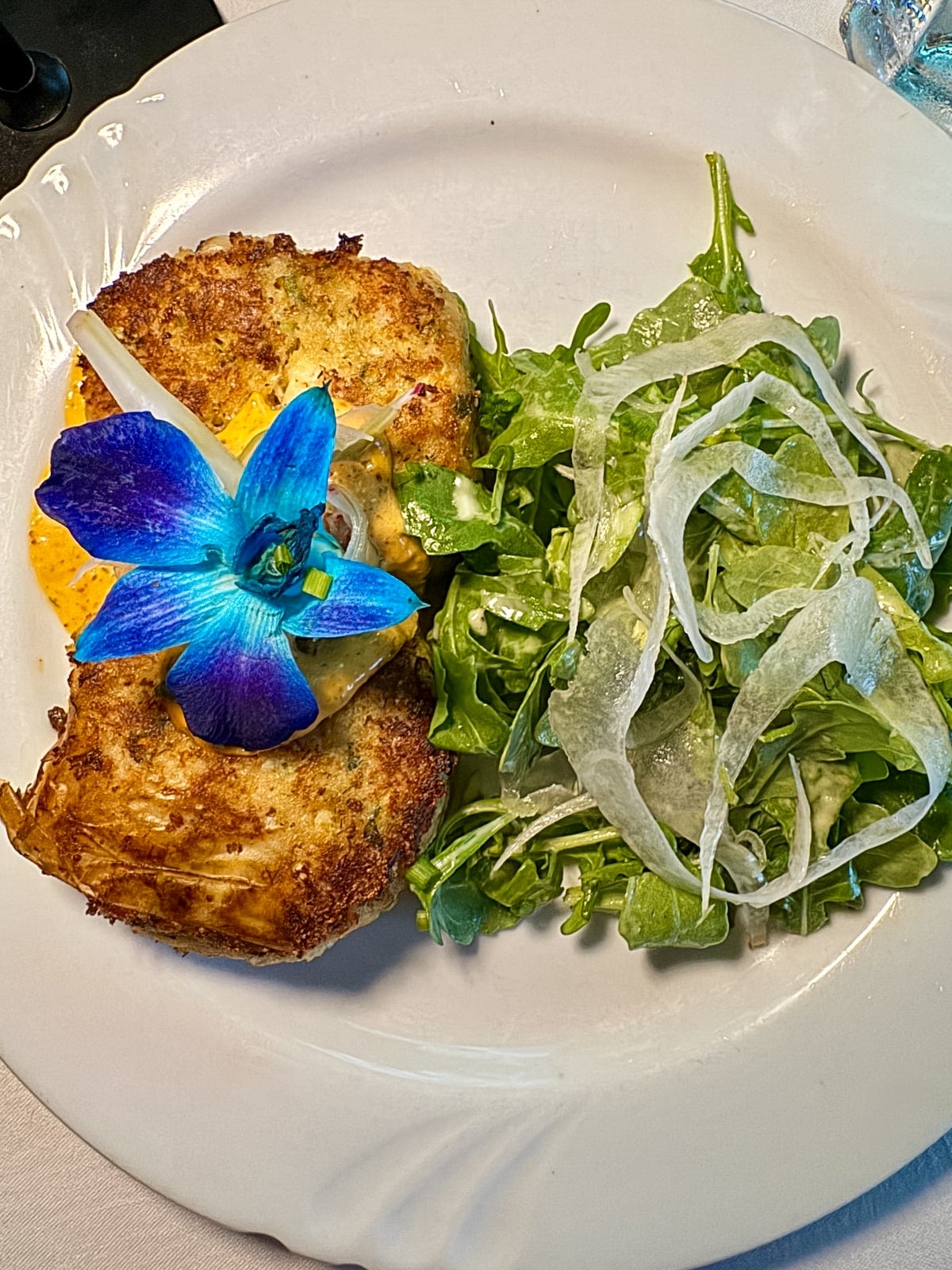 A crab cake garnished with a blue edible flower and creamy sauce, served with an arugula and shaved fennel salad on a white plate.
