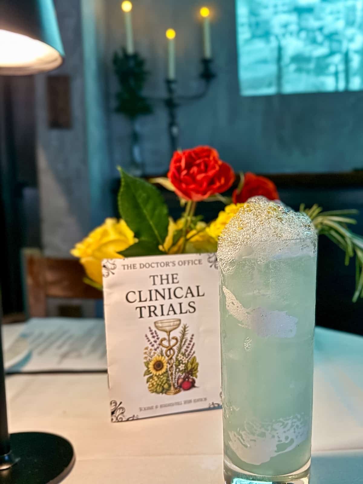 A tall glass of a fizzy drink sits on a table next to a cocktail menu titled &ldquo;The Clinical Trials,&rdquo; with a bouquet of red and yellow roses and candles in the background.