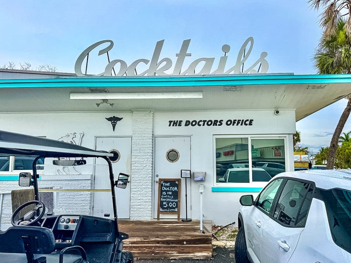 White building labeled &ldquo;The Doctors Office&rdquo; with a large &ldquo;Cocktails&rdquo; sign on the roof, a chalkboard sign by the door, and two cars parked in front.