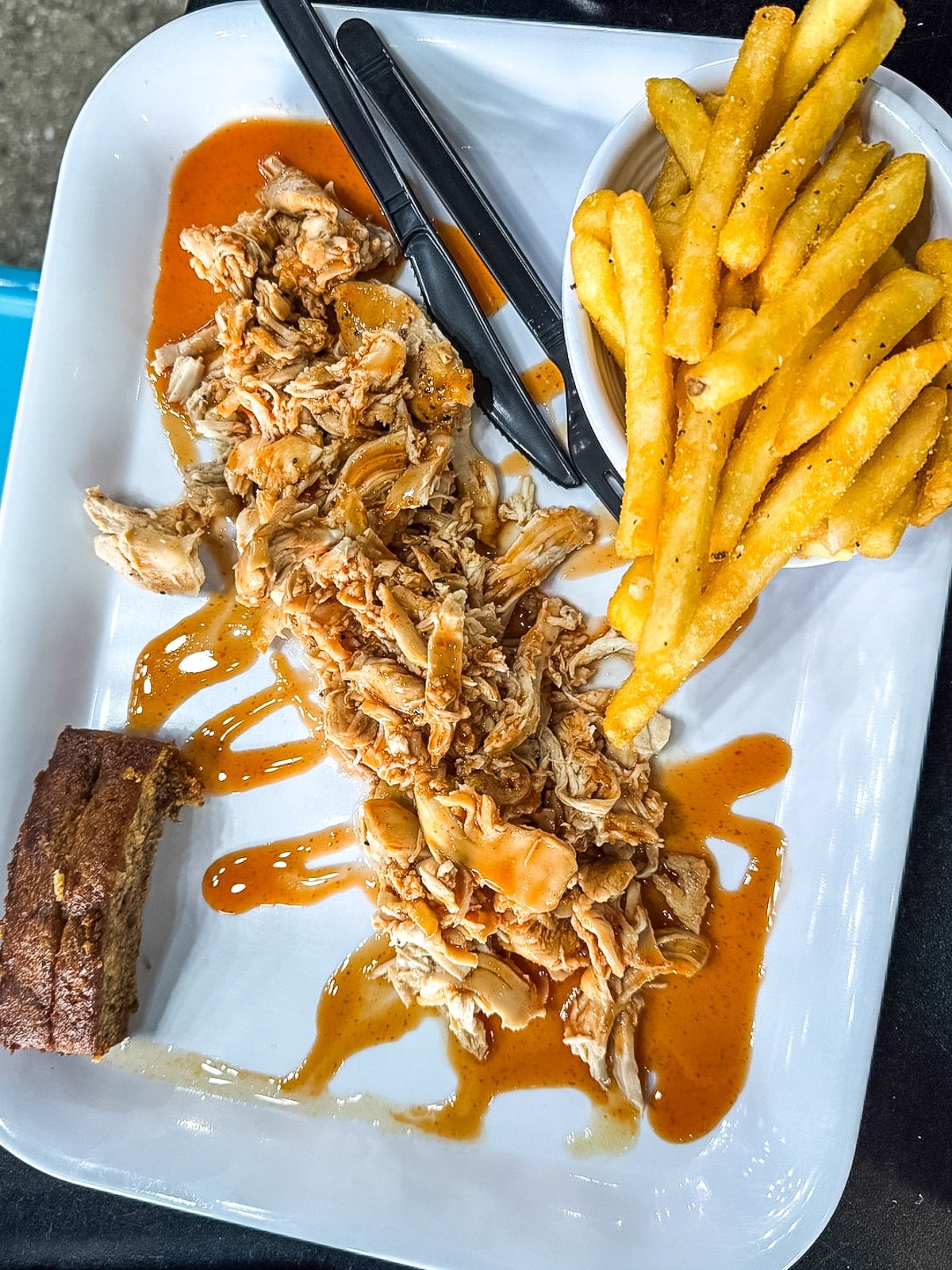 A plate with shredded barbecue chicken, French fries, a piece of cornbread, and black plastic cutlery on a white tray.