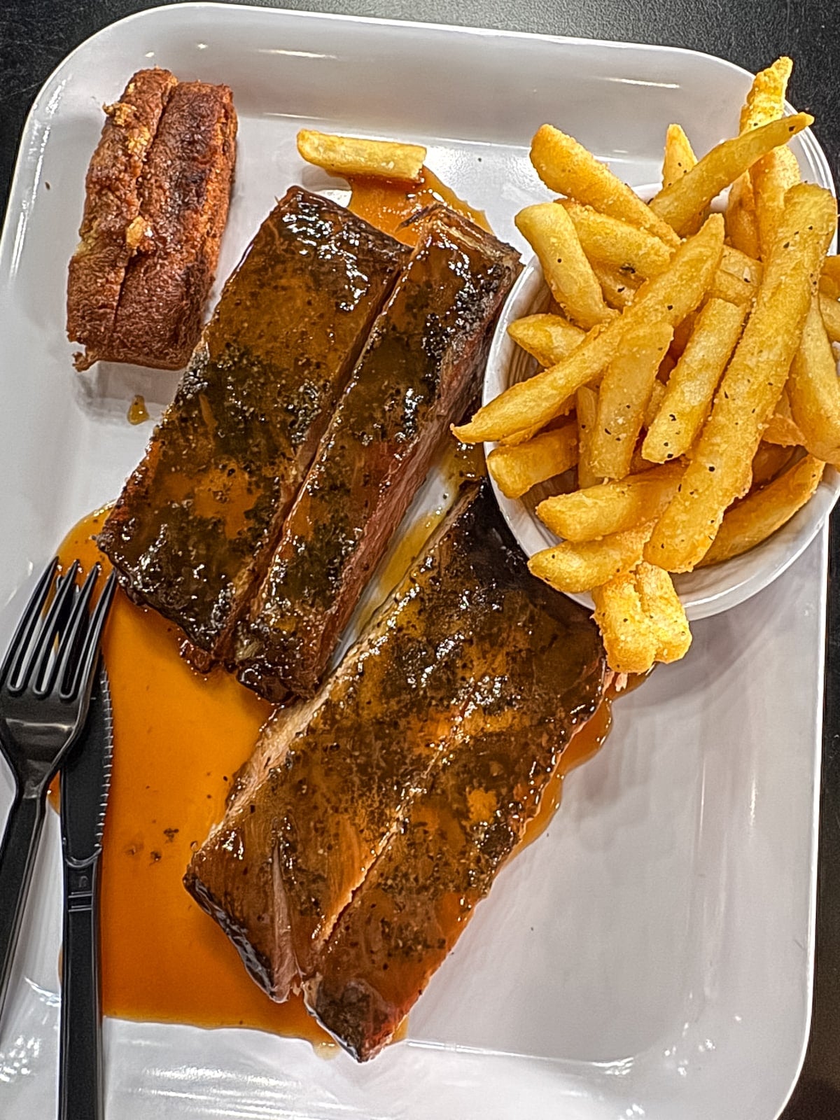 A plate with two glazed ribs, a serving of seasoned French fries in a bowl, a slice of cornbread, and plastic cutlery on the side.