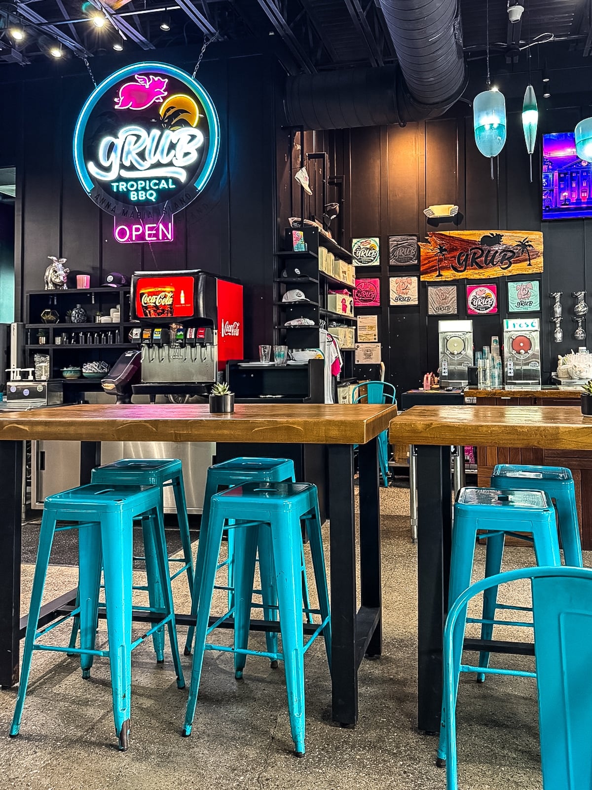 Interior of a casual restaurant with turquoise bar stools, wooden tables, a soda fountain, and neon signs on the wall. Shelves display various decor items and kitchen equipment.