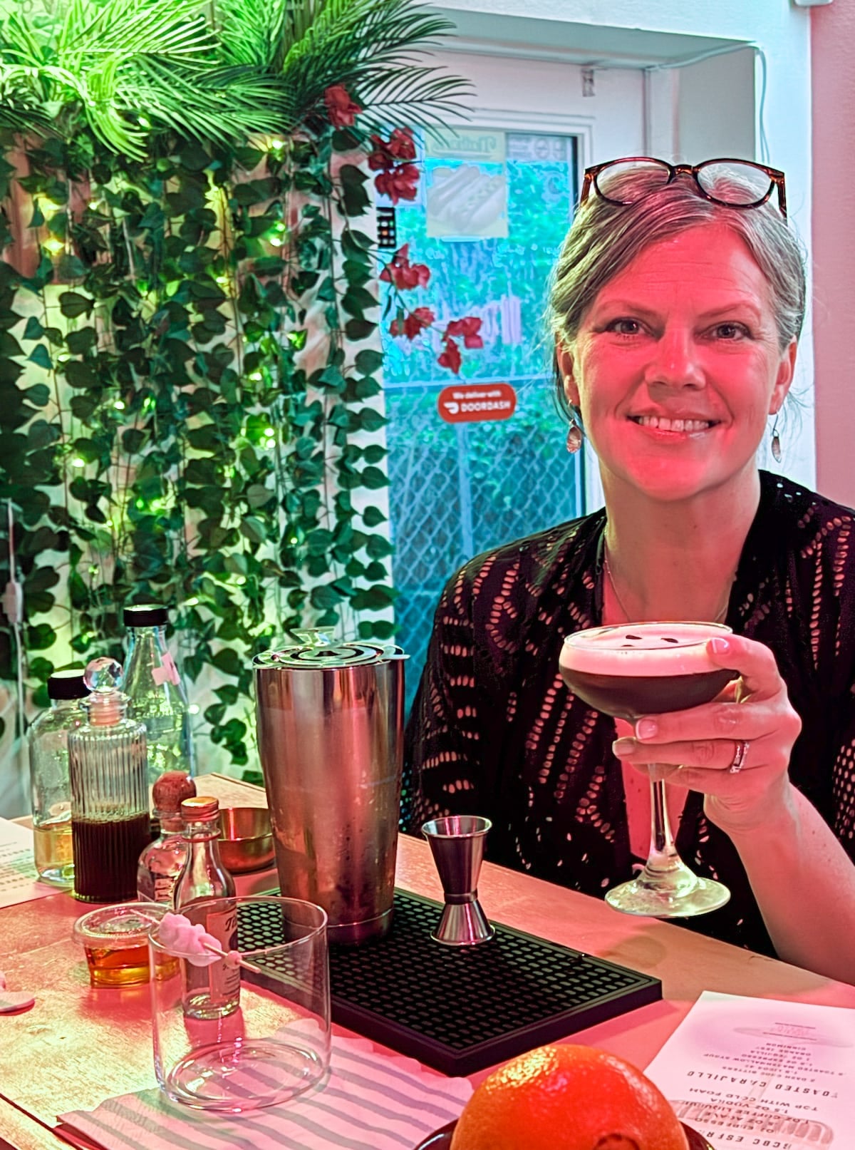 A woman in a black top holds a cocktail and smiles at a bar counter with various drink ingredients and tools under colorful lighting.