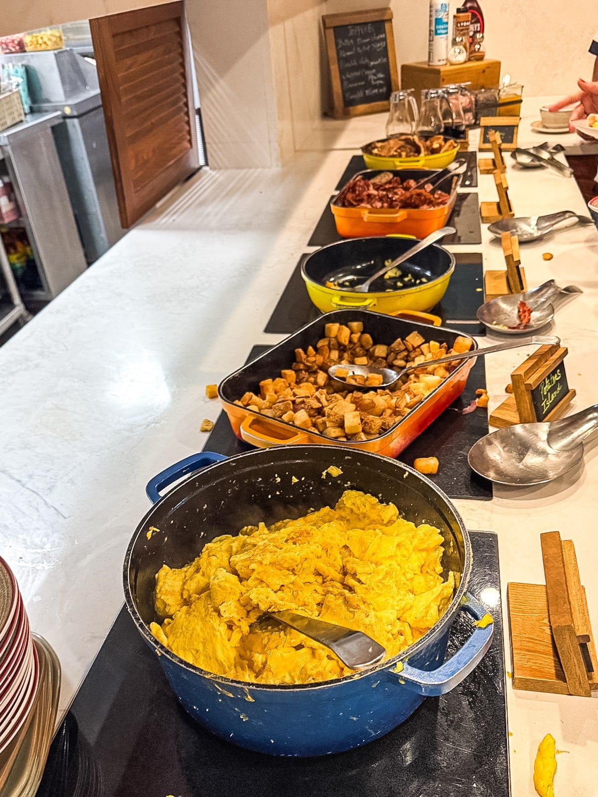 A breakfast buffet with scrambled eggs, potatoes, and other side dishes in serving trays on a counter with serving spoons.