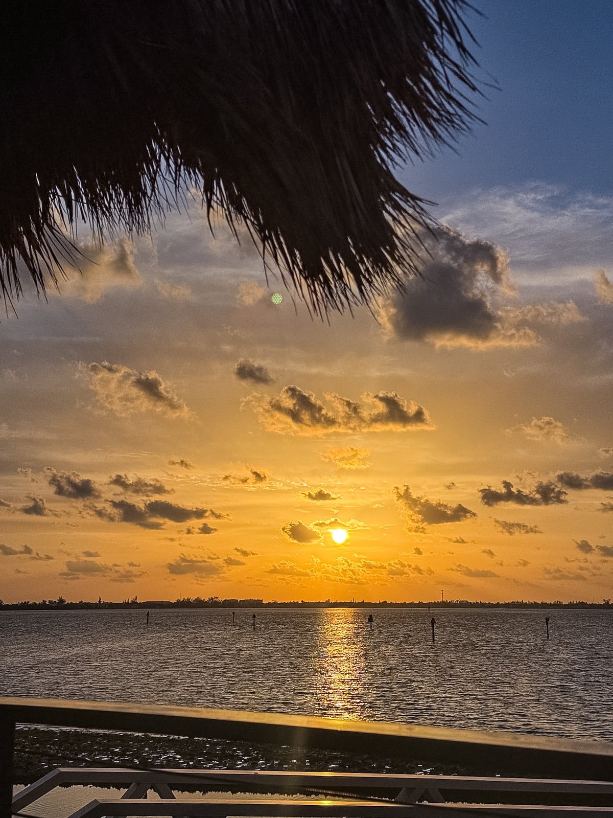 Sunset over a calm body of water with scattered clouds; a thatched roof is partially visible in the upper left corner.