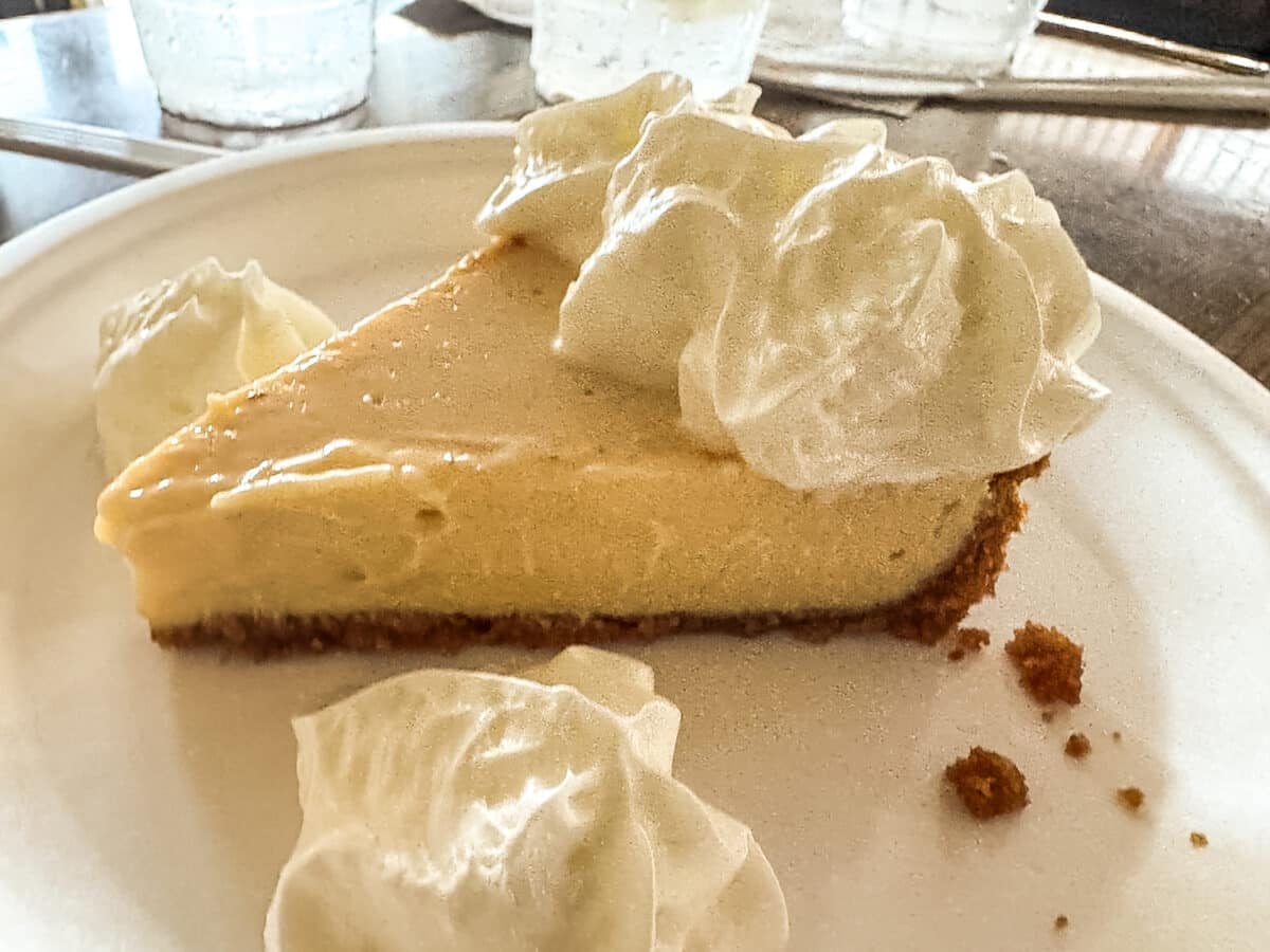 A slice of pie with a graham cracker crust is served on a plate, topped and surrounded by several dollops of whipped cream.