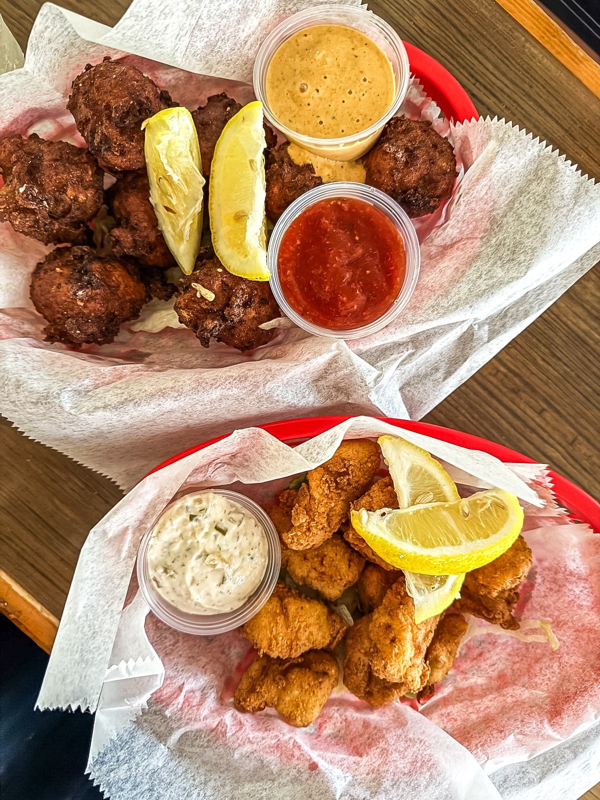 Two baskets of fried seafood and hush puppies with lemon wedges and dipping sauces, served on parchment paper in red plastic baskets on a wooden surface.