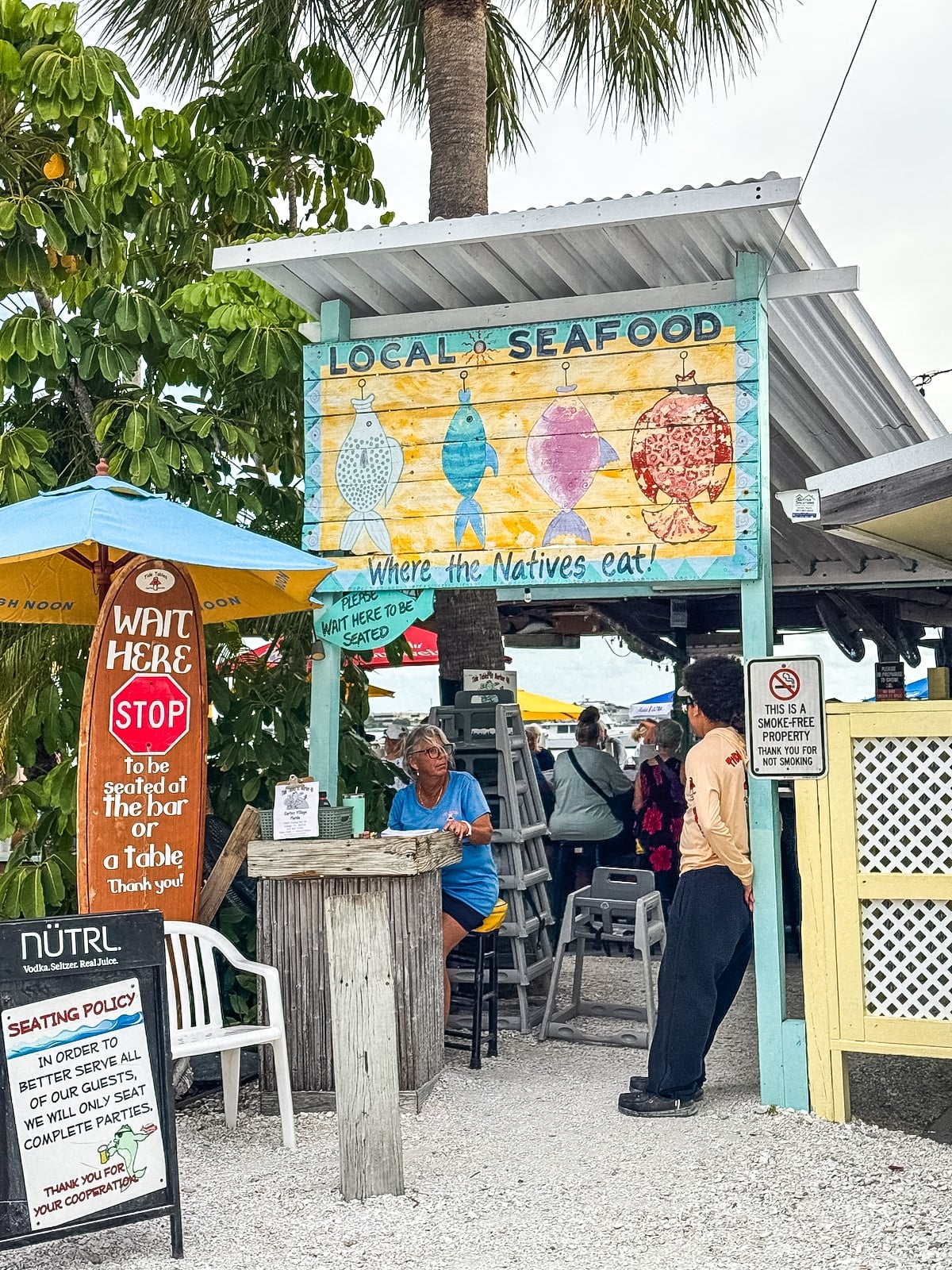 Outdoor seafood restaurant with a colorful &ldquo;Local Seafood: Where the Natives Eat&rdquo; sign, seating area, umbrellas, and various posted rules and seating policies.