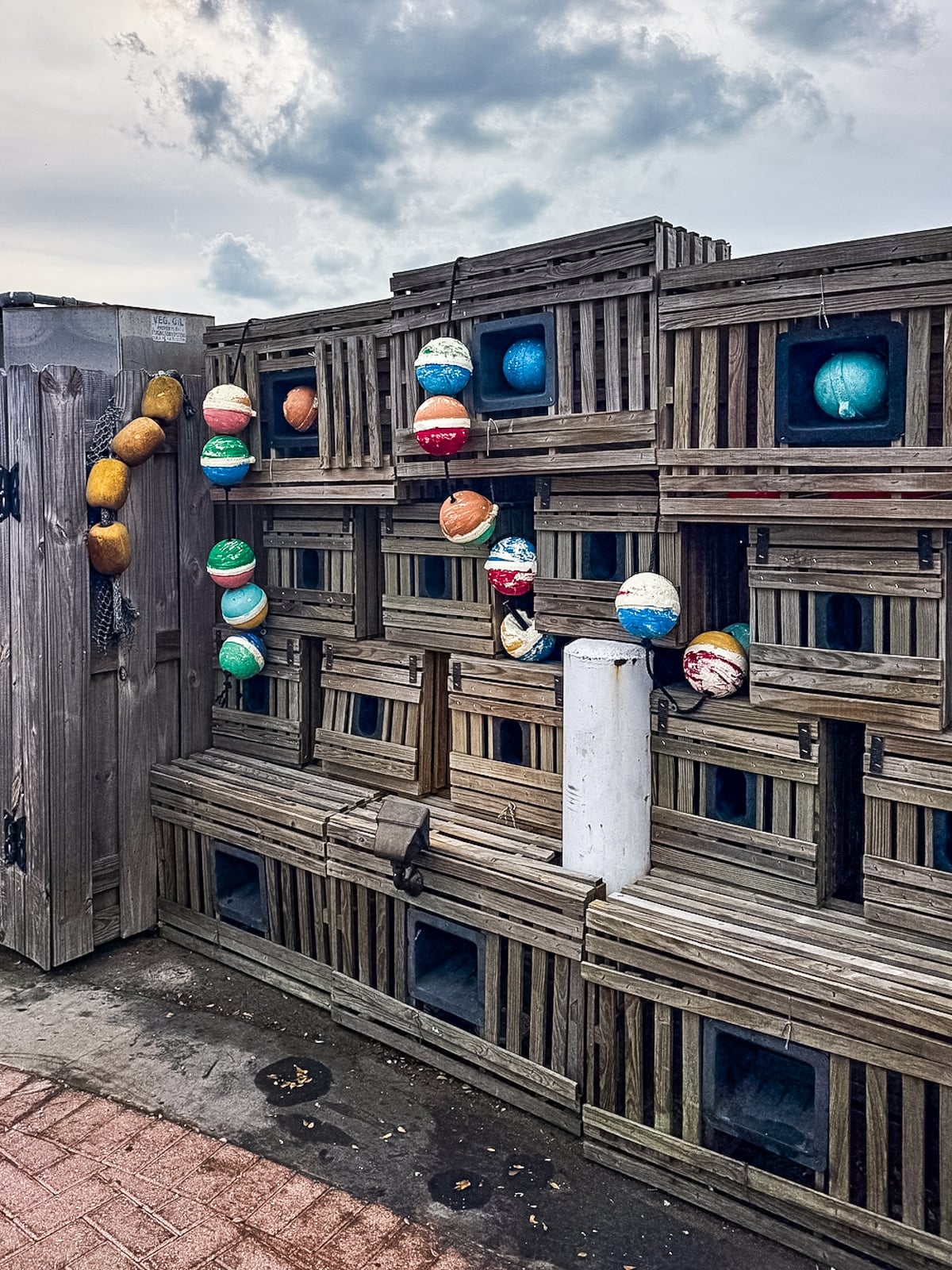 Stacked wooden lobster traps with colorful buoys hanging on the side, set outdoors on a cloudy day with a brick walkway in front.