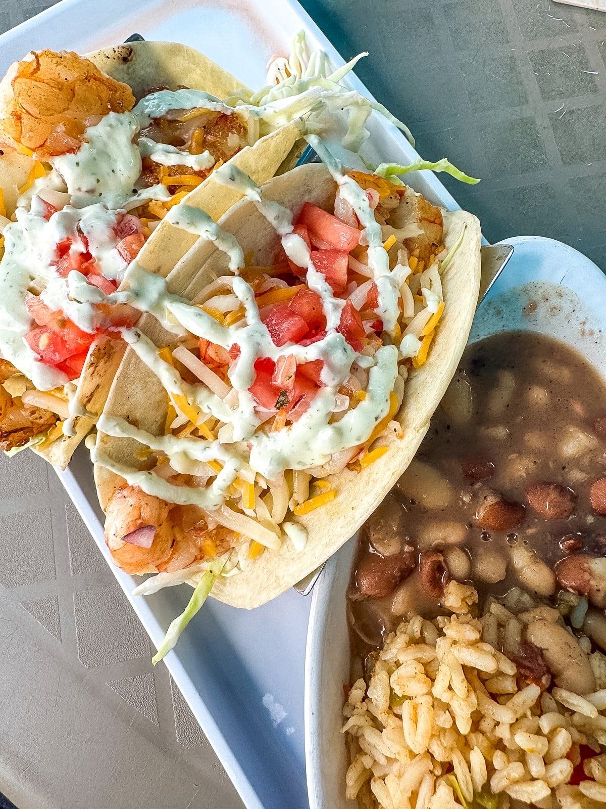 Three shrimp tacos with lettuce, tomato, cheese, and sauce are served on a plate next to a bowl of rice and beans.