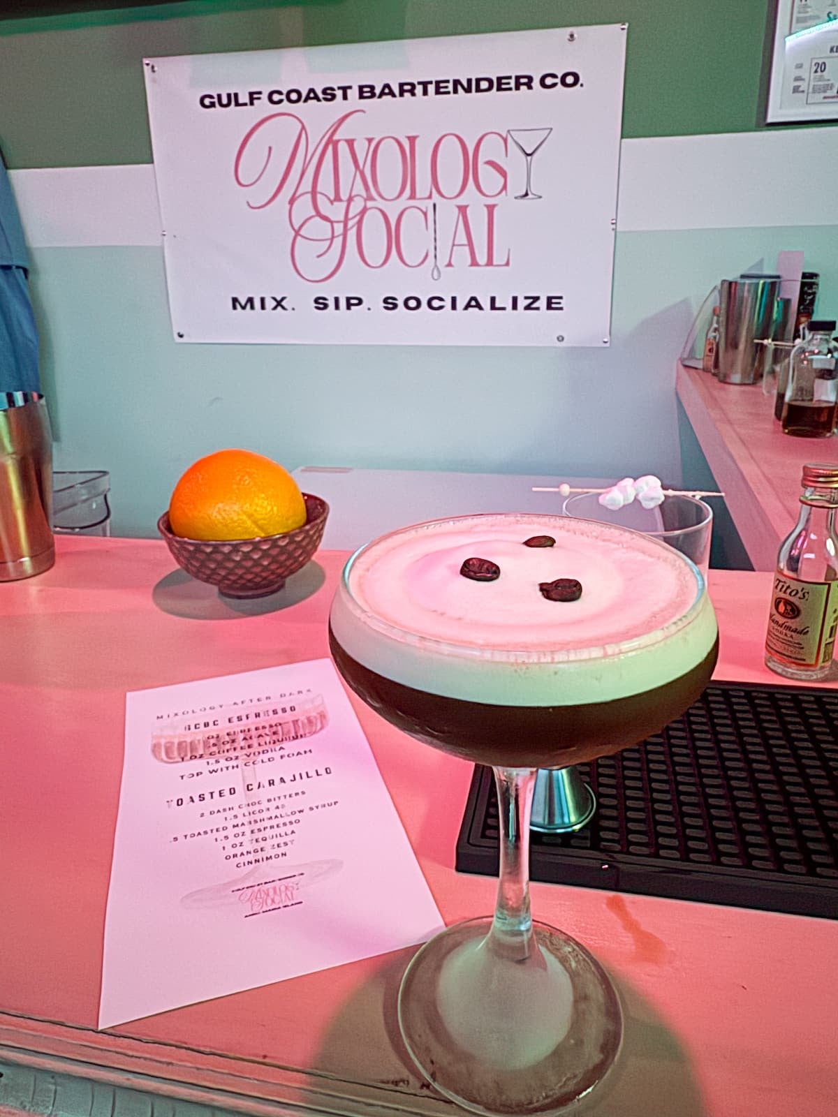A frothy cocktail garnished with three coffee beans sits on a bar counter next to an orange, a small menu, and bar tools under a &ldquo;Mixology Social&rdquo; sign.