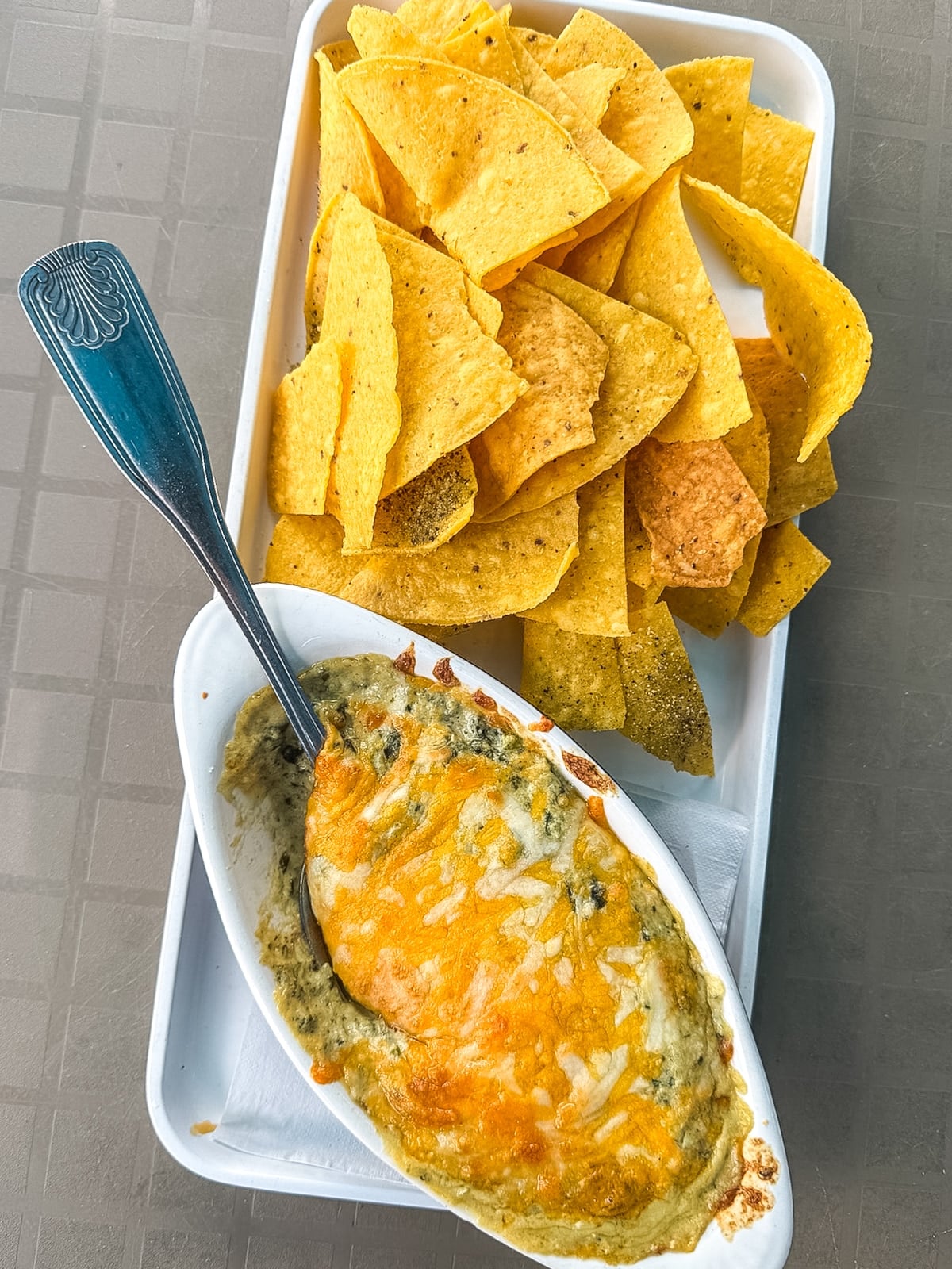 A tray with yellow tortilla chips and a small dish of baked cheese spinach dip, partially scooped, with a metal spoon.