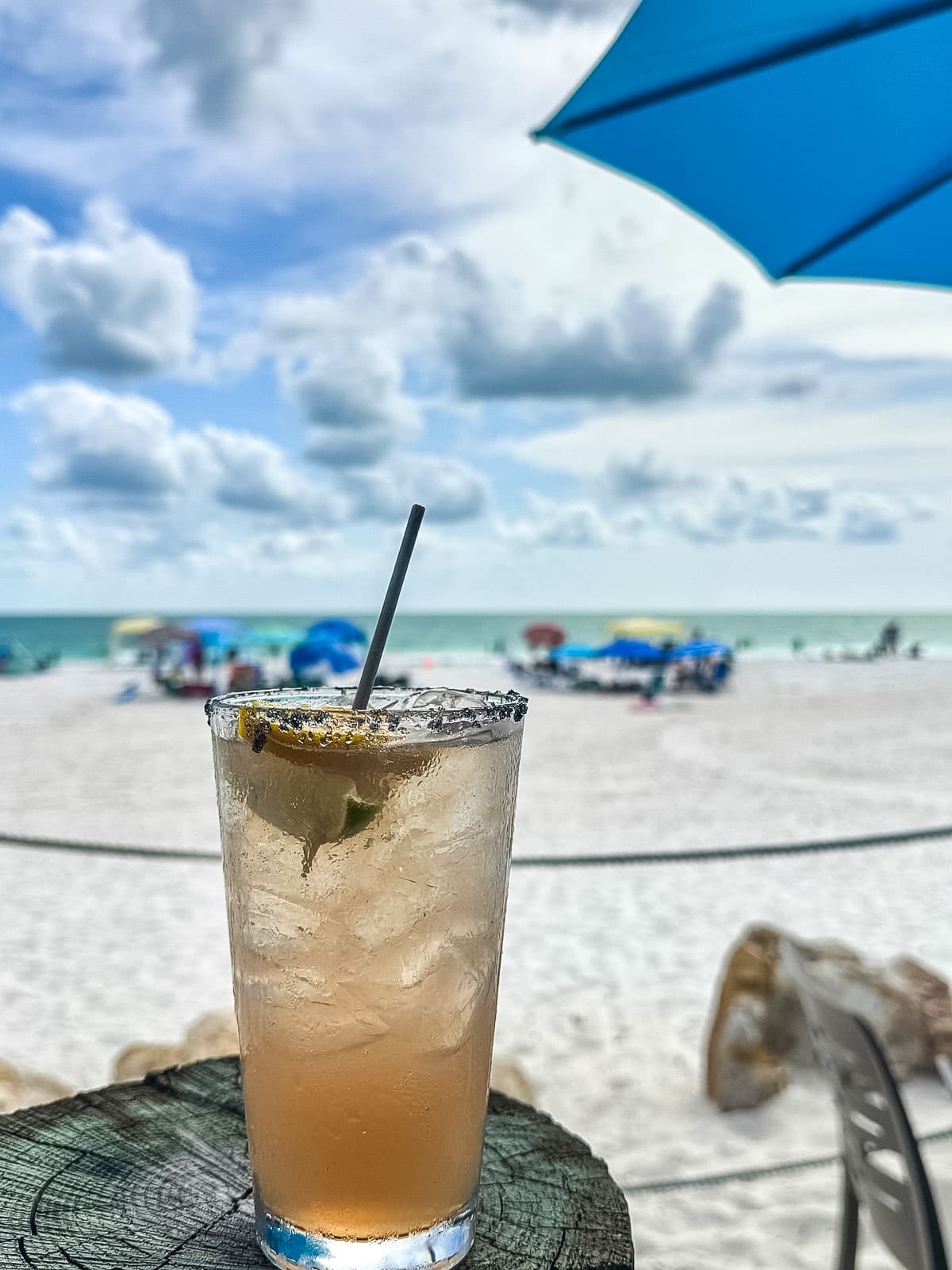 A cold drink with a straw and lime sits on a wooden surface. The background shows a sandy beach, blue ocean, umbrellas, and a partly cloudy sky.