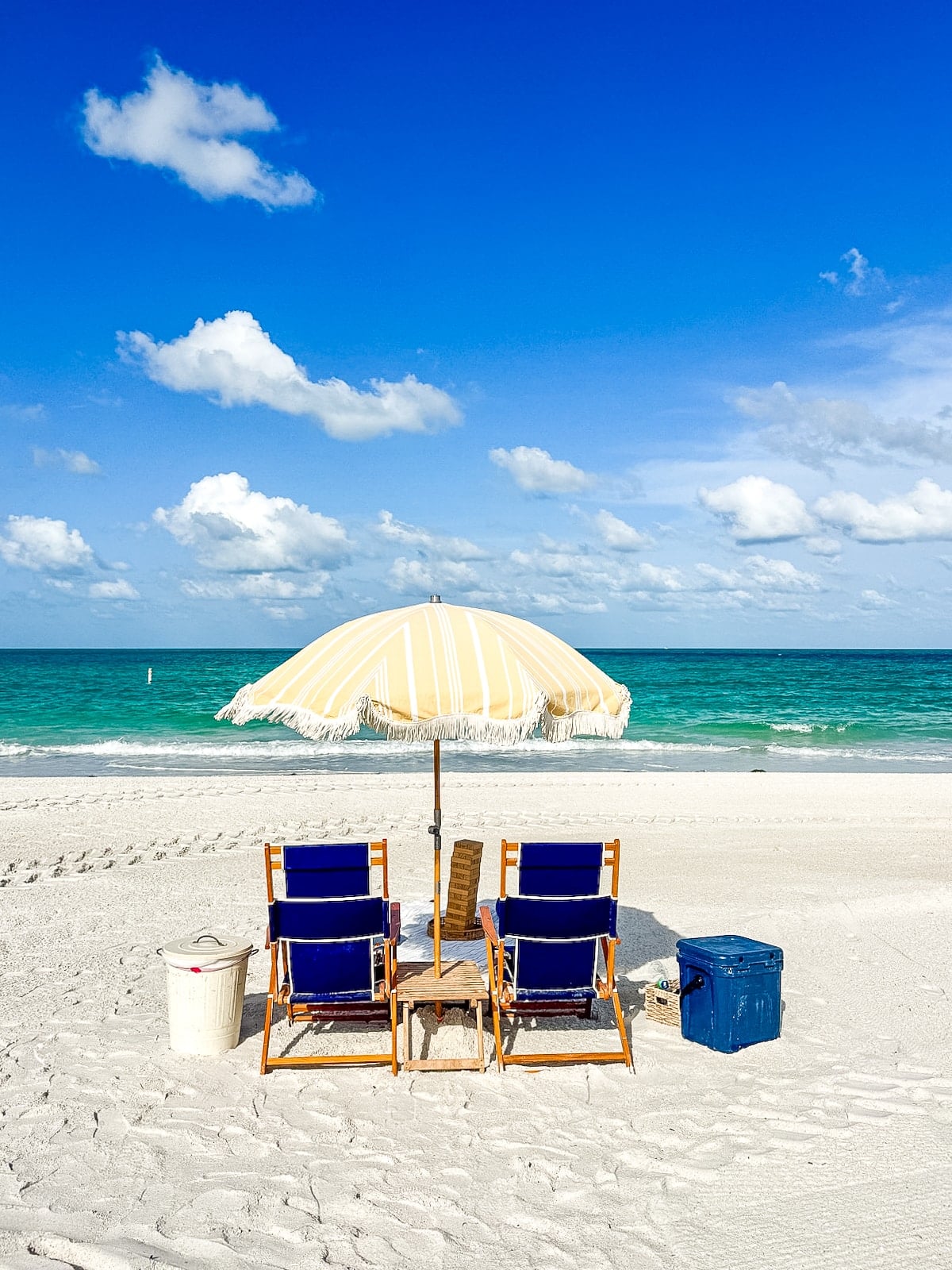 Two blue beach chairs and a striped umbrella face the ocean on white sand, with a bucket and cooler nearby under a bright blue sky.
