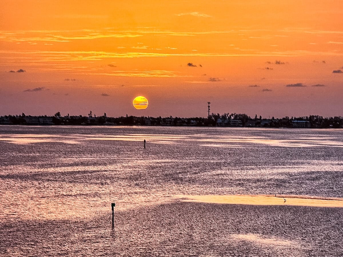 Orange sunset over calm water with the sun near the horizon, silhouetted land and trees in the background, and scattered clouds in the sky.
