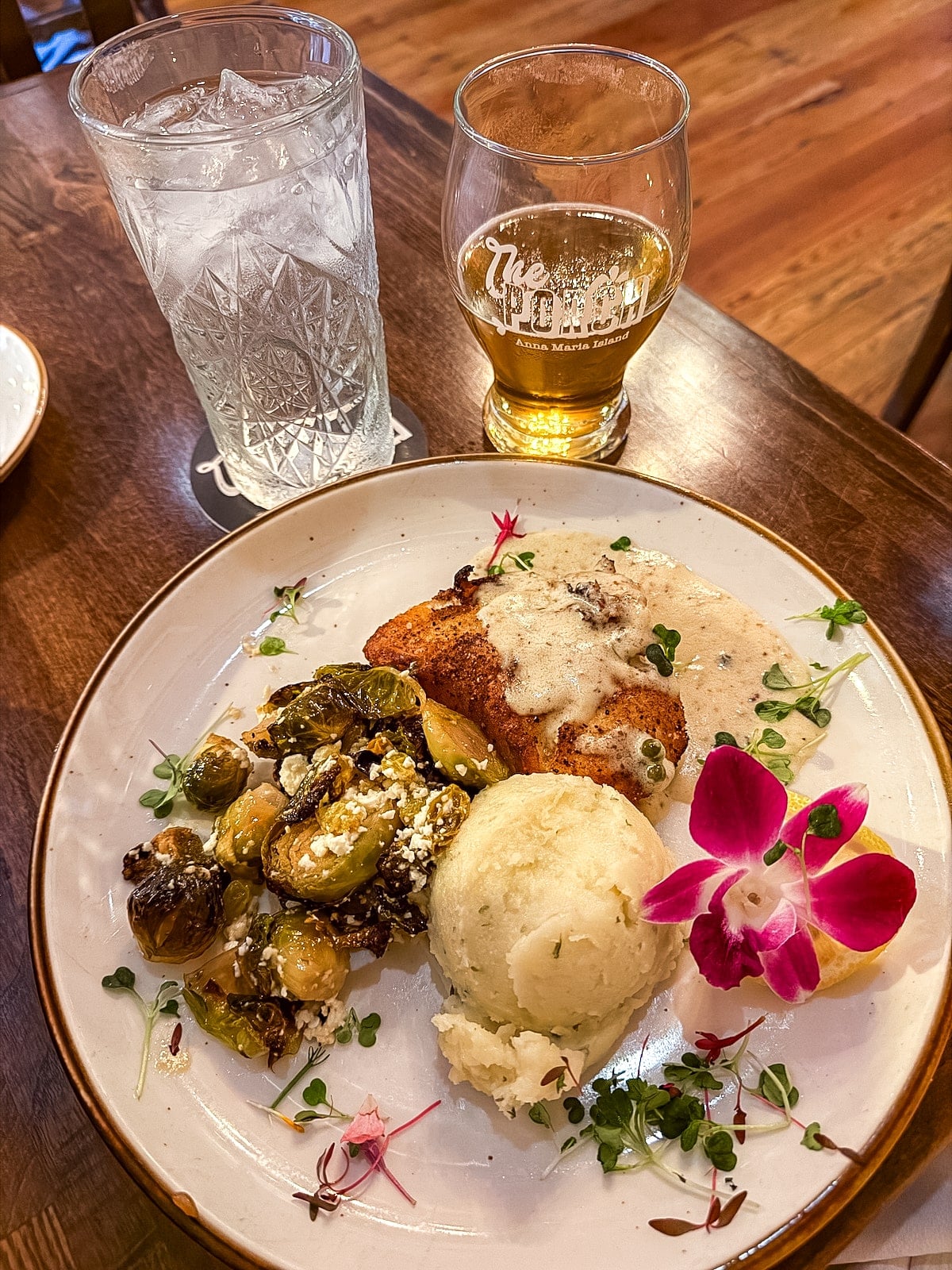 A plate with grilled fish, mashed potatoes, roasted Brussels sprouts, and edible flowers, served with a glass of water and a glass of beer on a wooden table.