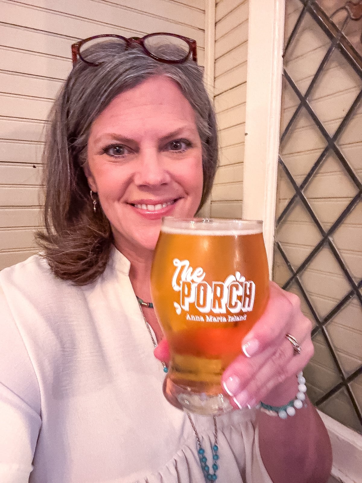 Woman with gray hair holds a glass of beer that reads &ldquo;The Porch, Anna Maria Island&rdquo; while smiling at the camera indoors.