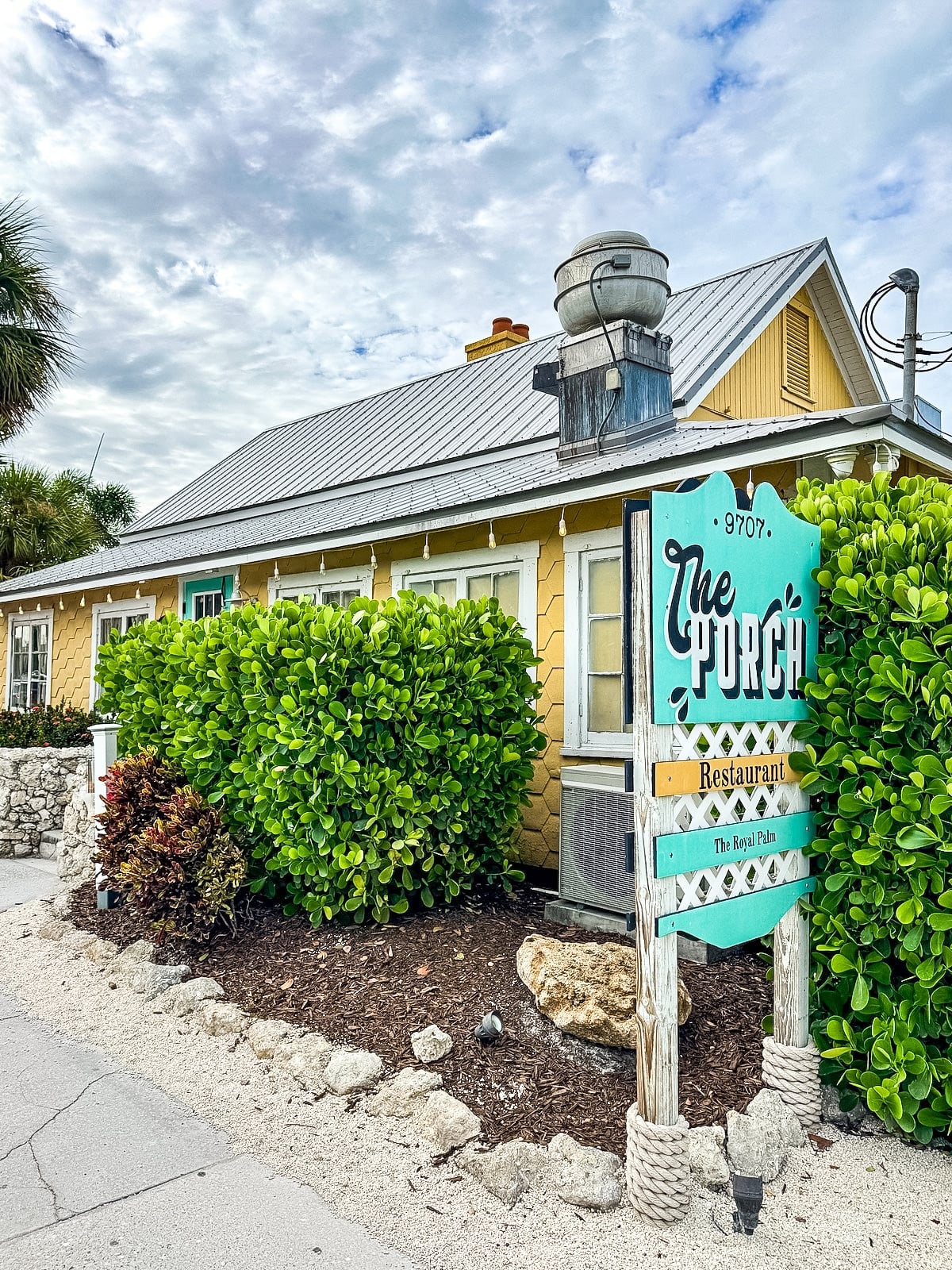 Yellow building with white trim, green bushes, and a turquoise sign reading &ldquo;The Porch Restaurant&rdquo; by a sidewalk under a cloudy sky.