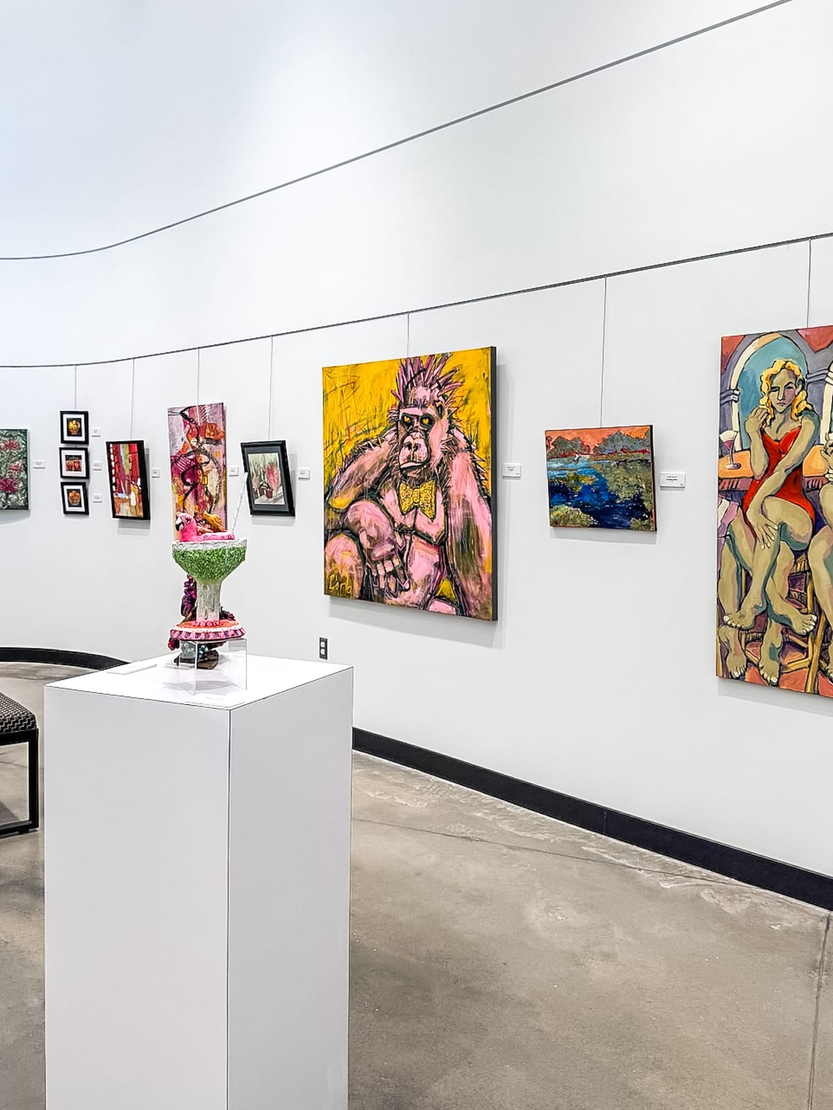 Art gallery with colorful paintings on white walls and a mixed-media sculpture displayed on a white pedestal in the foreground.