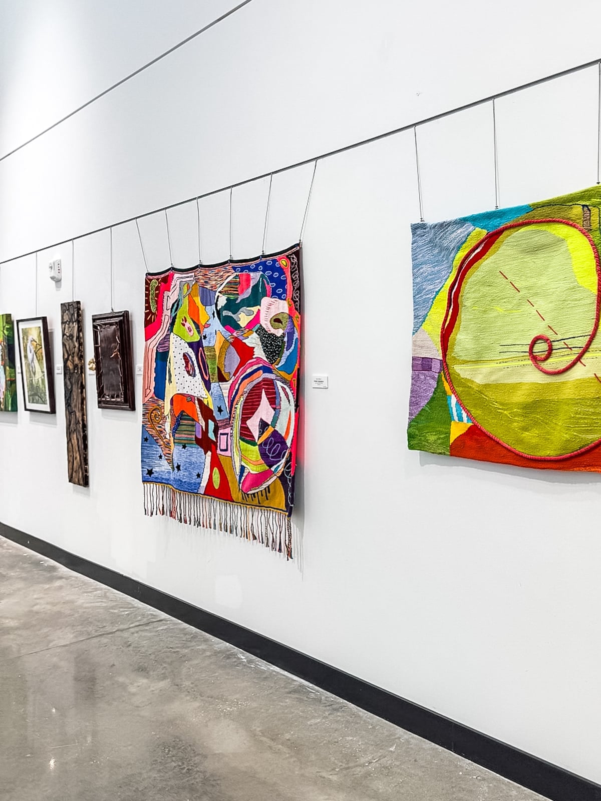 Artworks of varying styles and colors hang on a white gallery wall above a polished concrete floor.