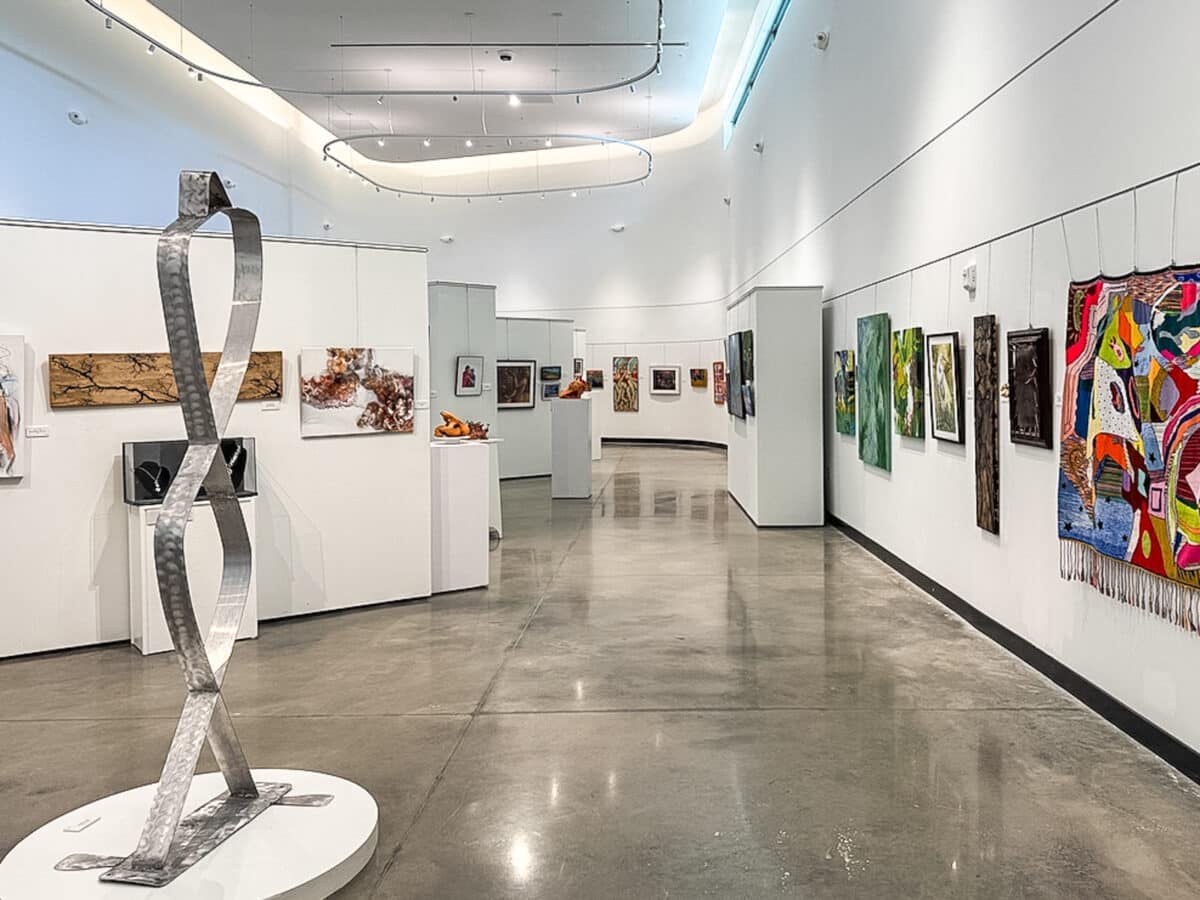 A modern art gallery with white walls, various colorful paintings, sculptures, and woven art displayed throughout the well-lit, polished space.