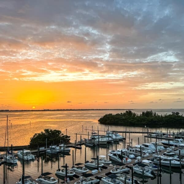 Boats docked in a marina at sunset, with the sun low on the horizon and clouds scattered across the sky over calm water.