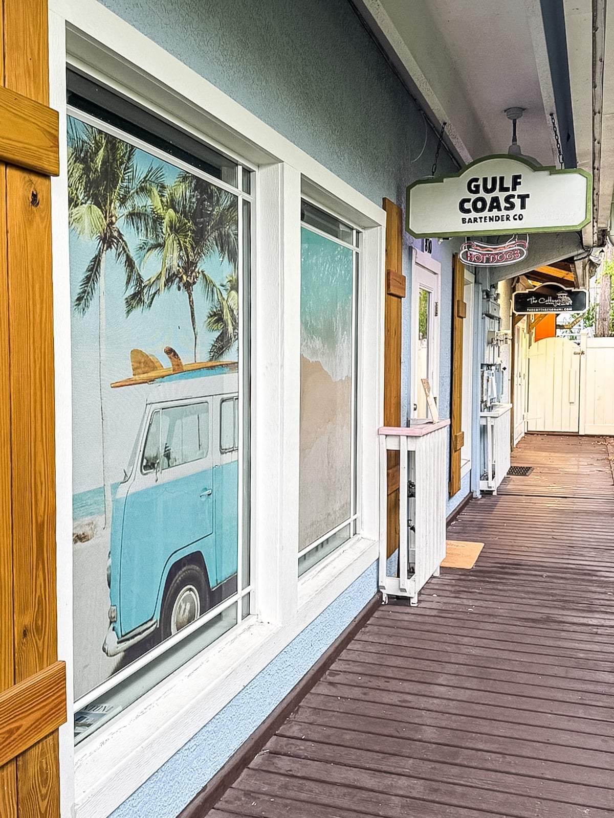 A boardwalk with a storefront, window art of a blue van under palm trees, and a sign reading &ldquo;Gulf Coast Barter Co&rdquo; hanging above the walkway.