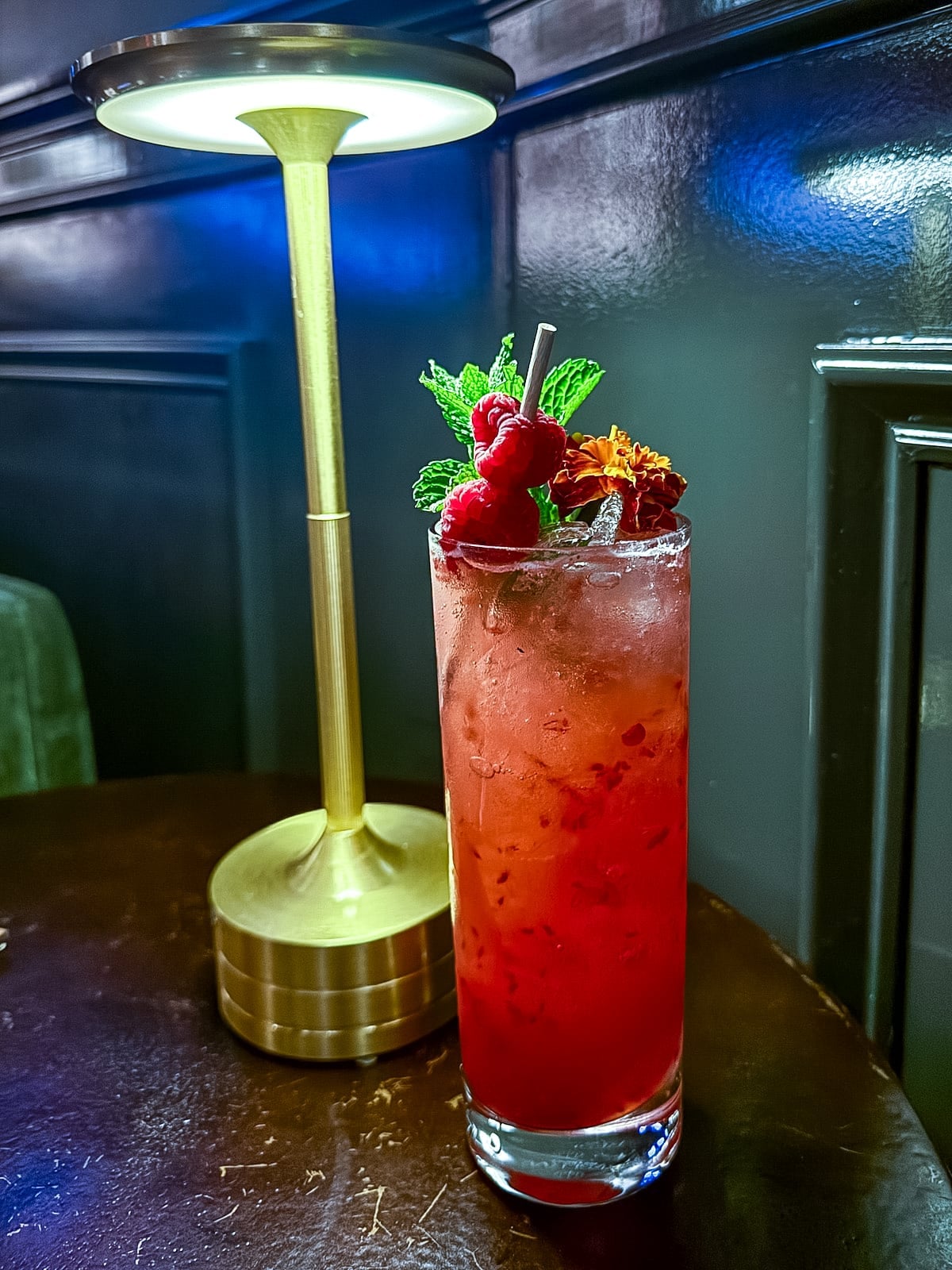 A tall glass of red berry cocktail with ice, mint, raspberries, and an orange flower garnish sits on a dark table next to a gold lamp.