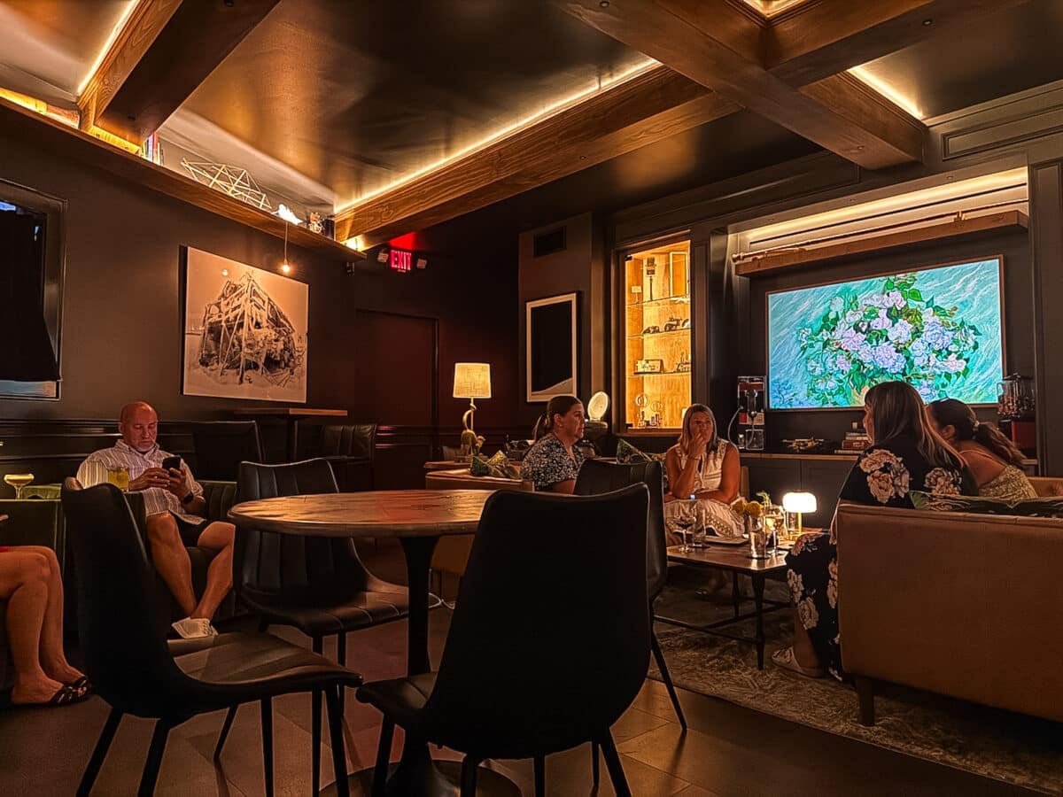 A dimly lit lounge with people sitting on sofas and chairs, chatting and using phones. A floral image is displayed on a screen, and artwork hangs on the wall.