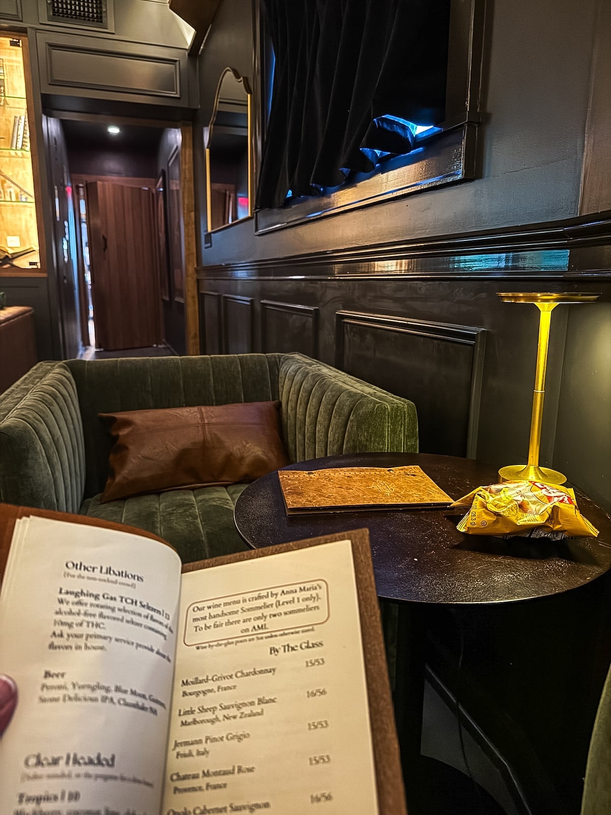 A person holds a menu in a dimly lit lounge with a green sofa, a small round table, a glowing lamp, and bags of chips.