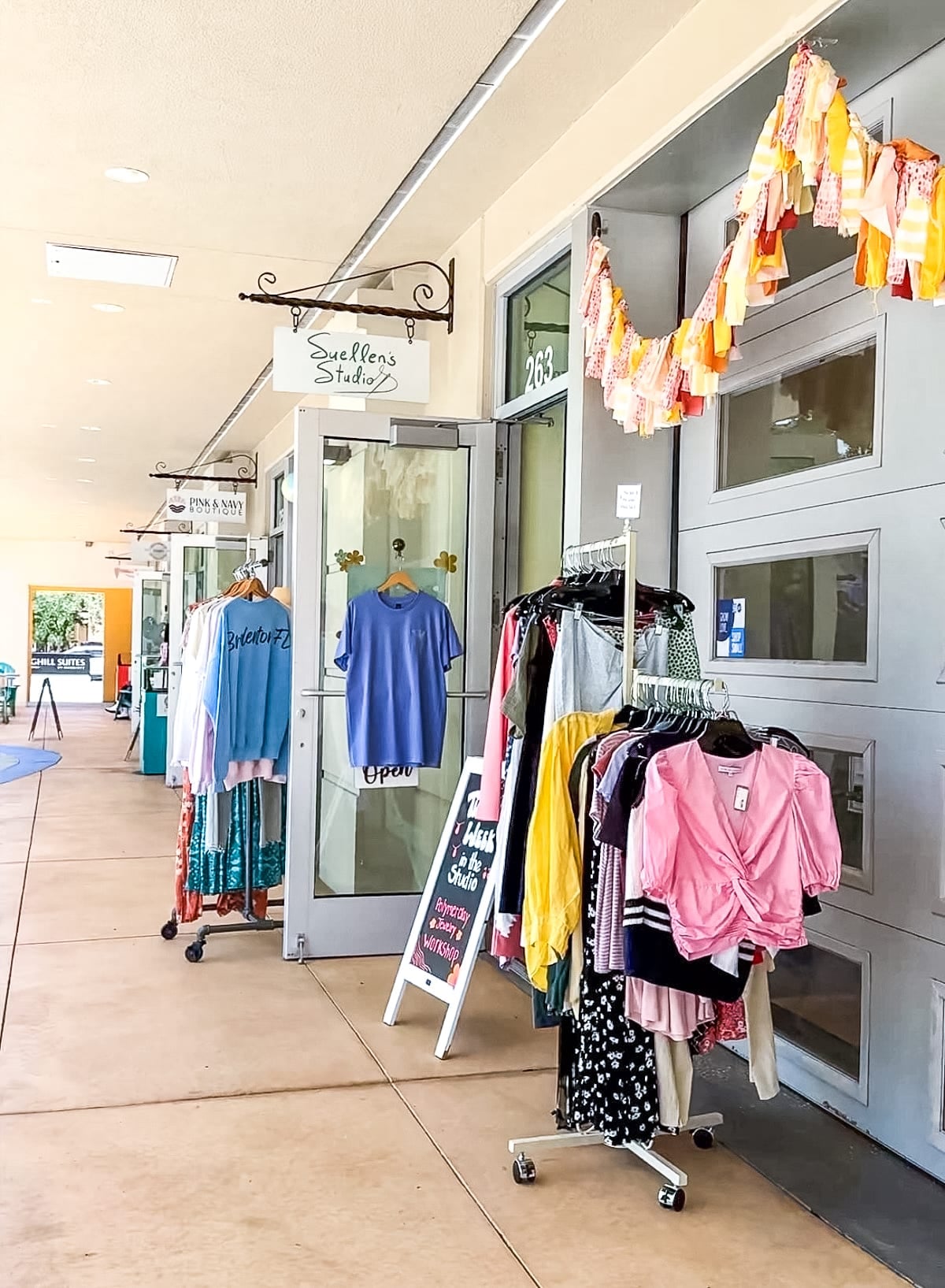 Outdoor shopping area with clothing racks, a chalkboard sign, and an open boutique door displaying shirts and dresses. Festive garland decorates the entry.