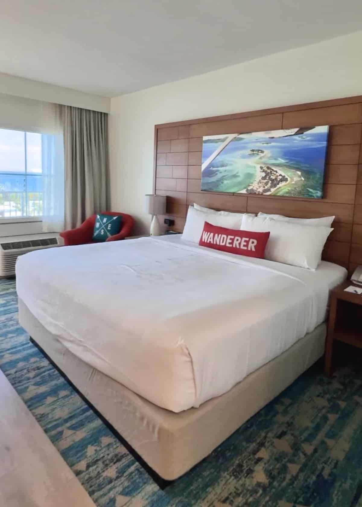 Hotel room with a large bed, red pillow that says &ldquo;WANDERER,&rdquo; red chair, window with ocean view, and a beach aerial photo above the headboard.